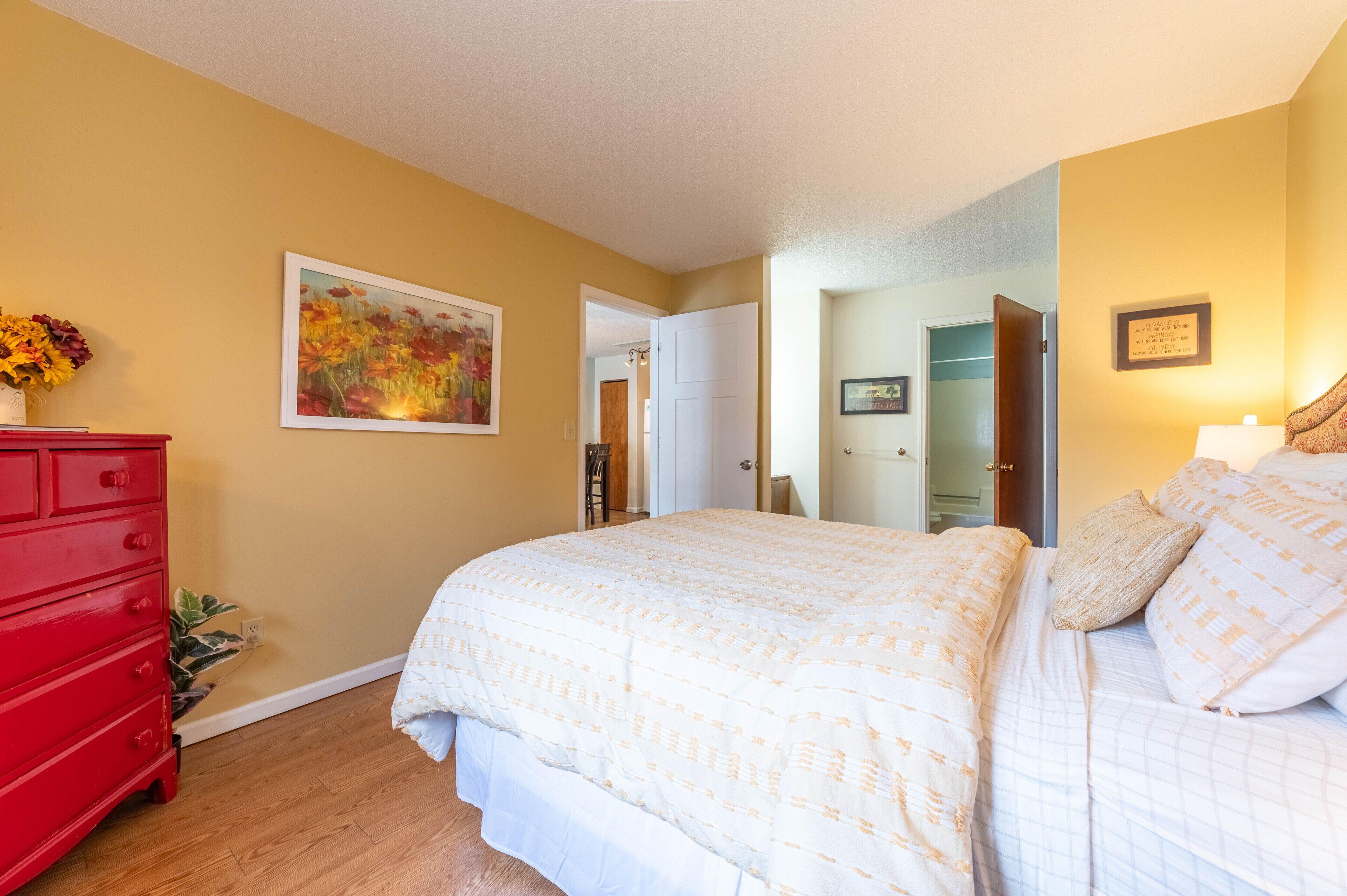 Un dormitorio con una cama grande, un tocador rojo, un cuadro en la pared y una puerta que da a otra habitación.