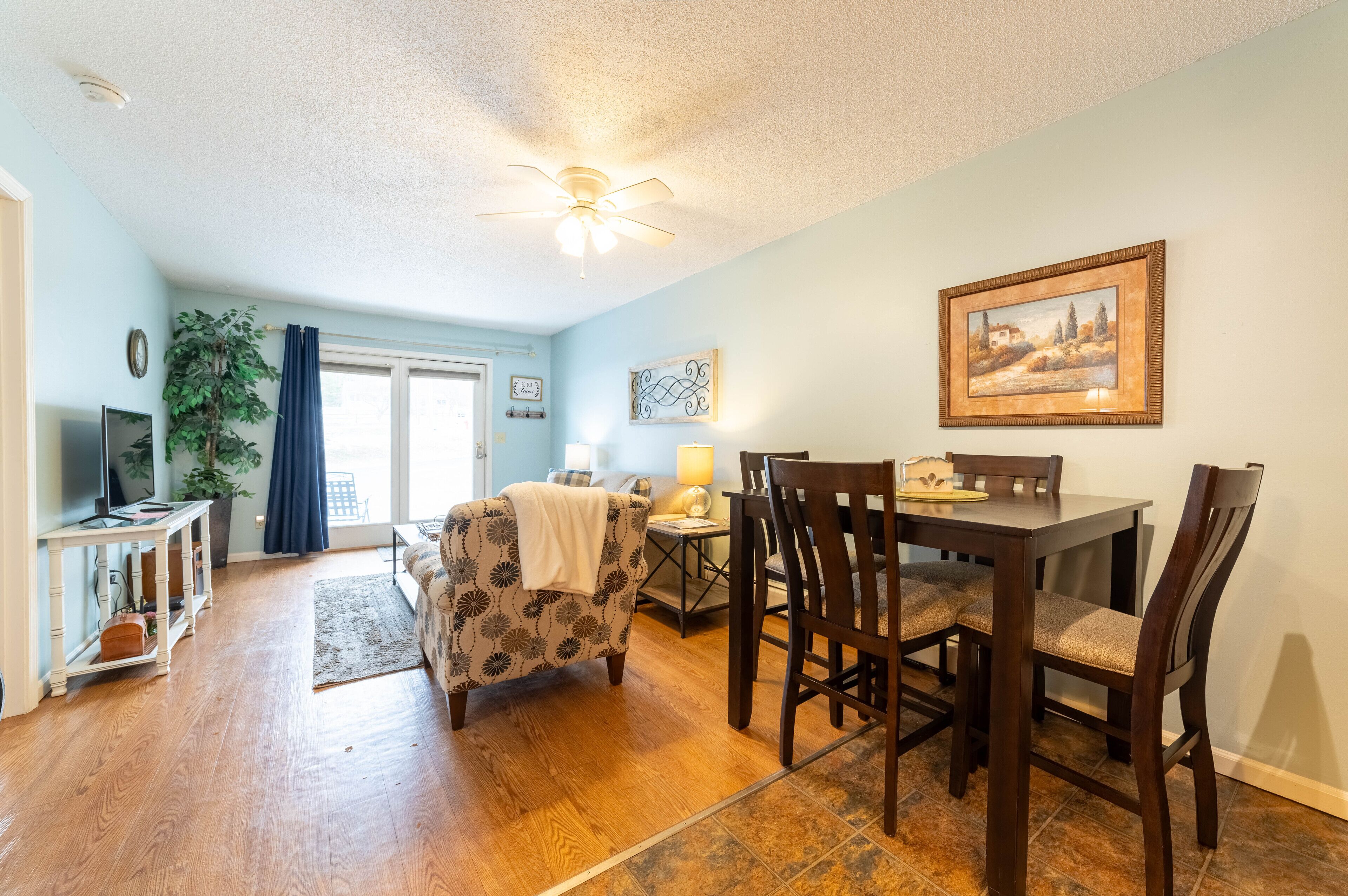 A dining area with a wooden table and chairs, a ceiling fan, and a painting on the wall.