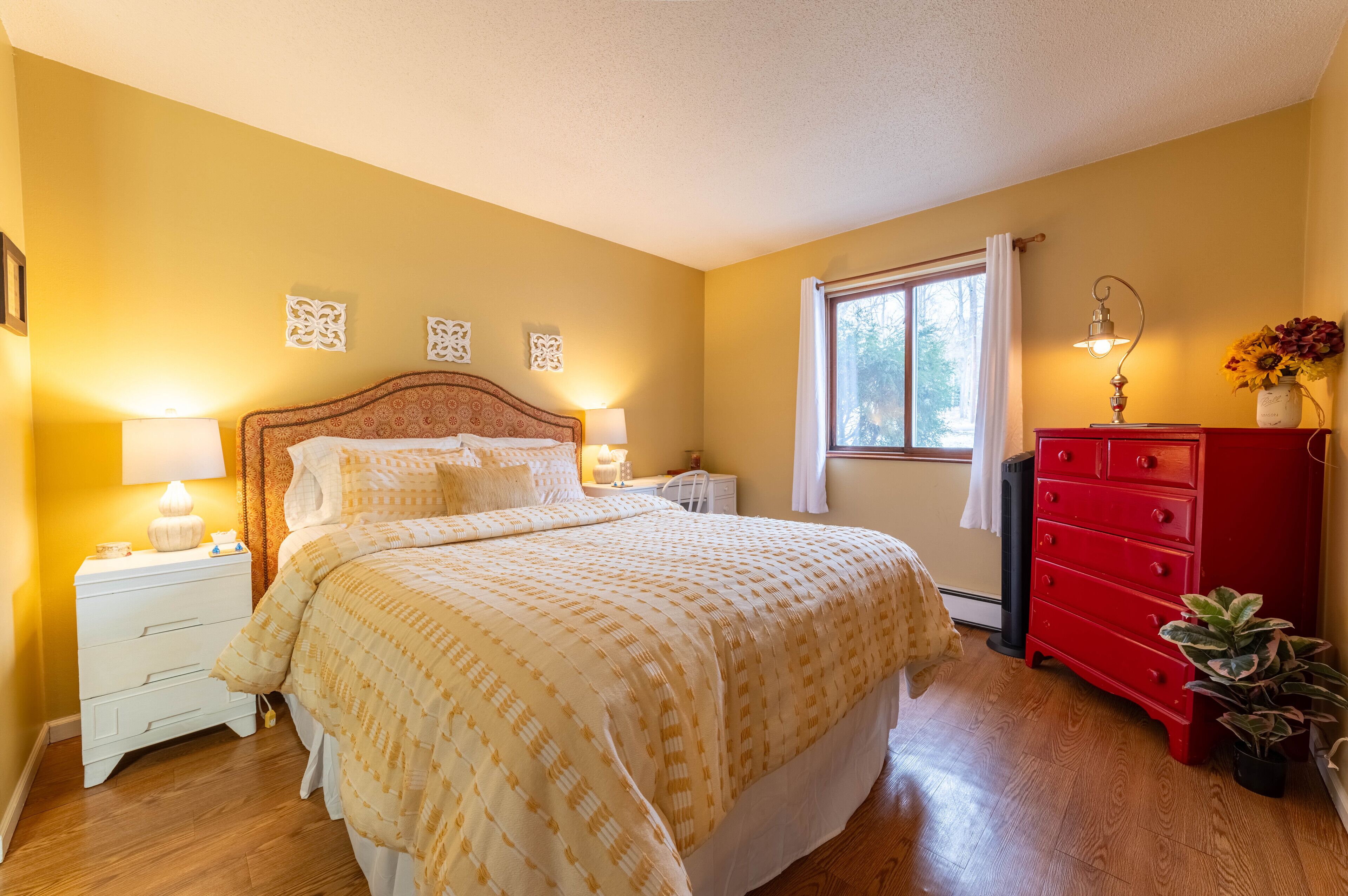 A bedroom with a large bed, two bedside tables with lamps, a red dresser, and a window with curtains.
