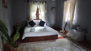 A bedroom with a wooden bed, white bedding, two dark pillows, a floral rug, a small bedside table with a kettle, and a potted plant.