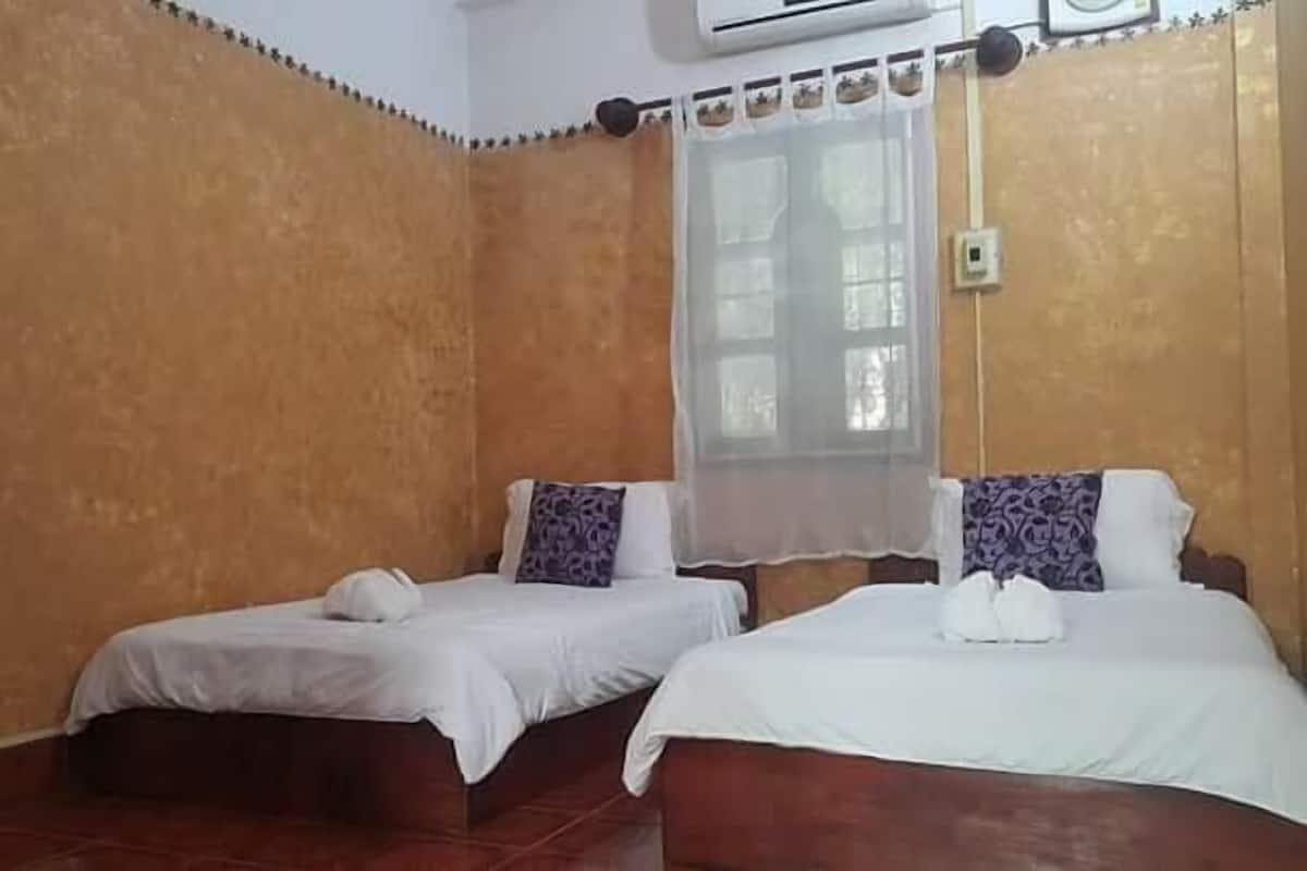 A room with two beds, a window with a sheer curtain, a wall-mounted air conditioner, and a small table with a vase.