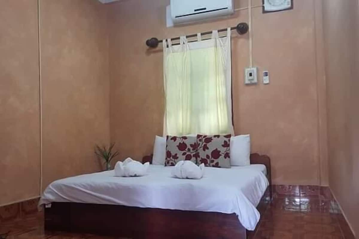 A bedroom with a bed, a small bedside table, a window with curtains, and an air conditioning unit on the wall.