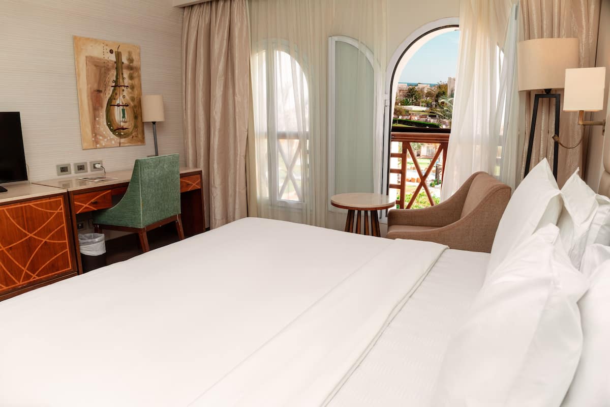 A hotel room with a large bed, a desk with a chair, a small table, and a view of the outdoors through a glass door.