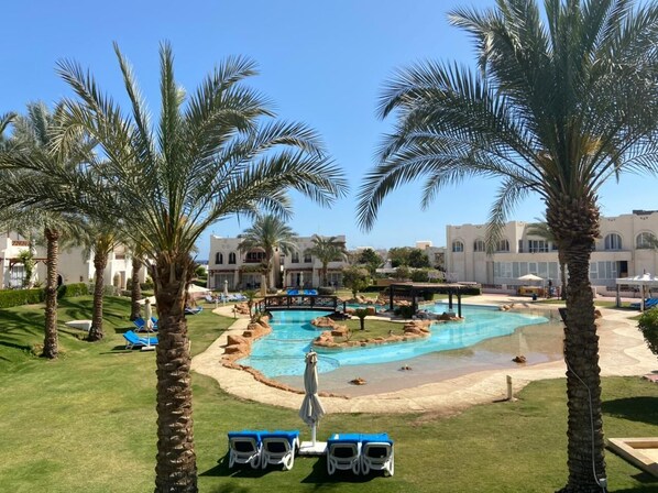 Zona de piscina en un resort, con palmeras, sillas de descanso y un cielo azul brillante.