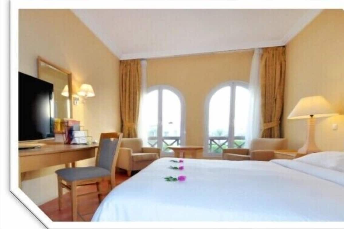 A hotel room with a bed, a desk with a chair, a TV, and two windows with curtains.