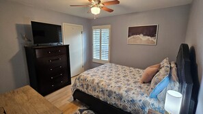 Een slaapkamer met een bed, een dressoir, een plafondventilator, een televisie op het dressoir en een schilderij aan de muur.