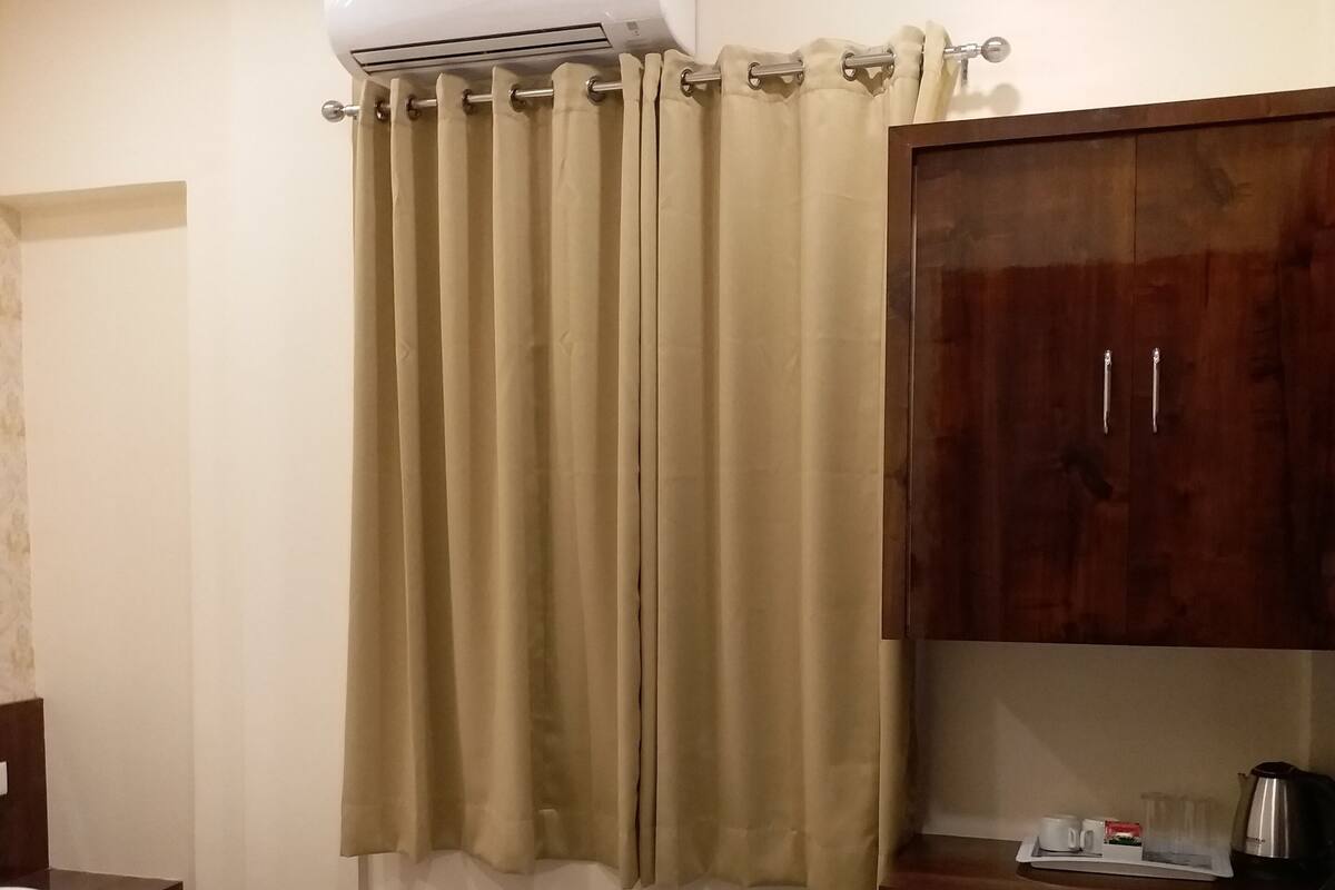 A hotel room with a bed, an air conditioning unit, a wooden cabinet, and a tray with a coffee pot and cups.