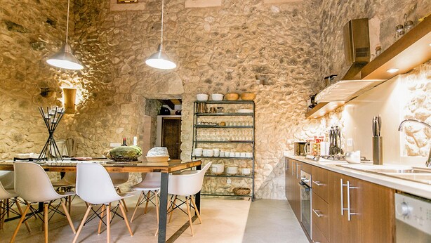 Un interior rústico de piedra con una mesa de comedor de madera, sillas blancas, luces colgantes, un estante empotrado con vajilla y una zona de cocina moderna.