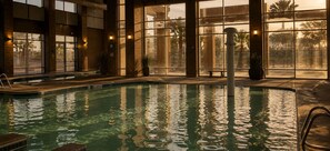 An indoor swimming pool with clear water, surrounded by large windows and potted plants.