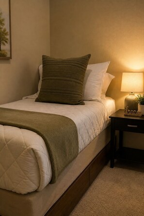 Un lit bien fait, recouvert d’une courtepointe matelassée, avec un plaid vert et un oreiller. On y trouve une table de chevet avec une lampe et une petite plante.