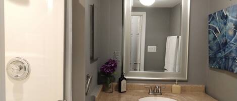 A bathroom with a white shower, a sink with a beige countertop, a dark wooden cabinet, a large mirror, and a purple flower vase.