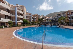 A swimming pool surrounded by multi-story buildings with balconies, a clear sky, and a mountainous backdrop.