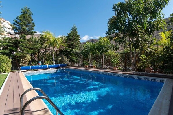 A clear blue pool surrounded by lush greenery and trees, with a wooden deck and a patterned pool edge.
