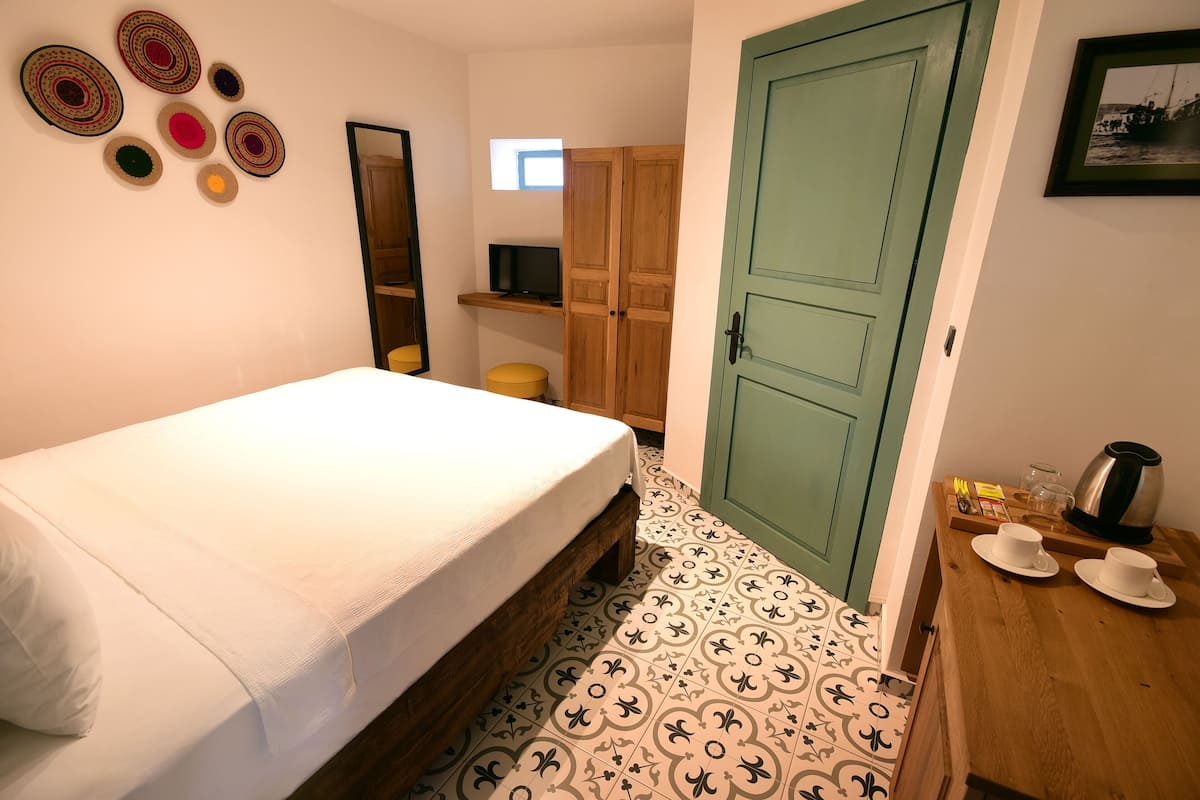 A bedroom with a bed, a wooden wardrobe, a small table with cups and a kettle, and a wall with decorative plates.