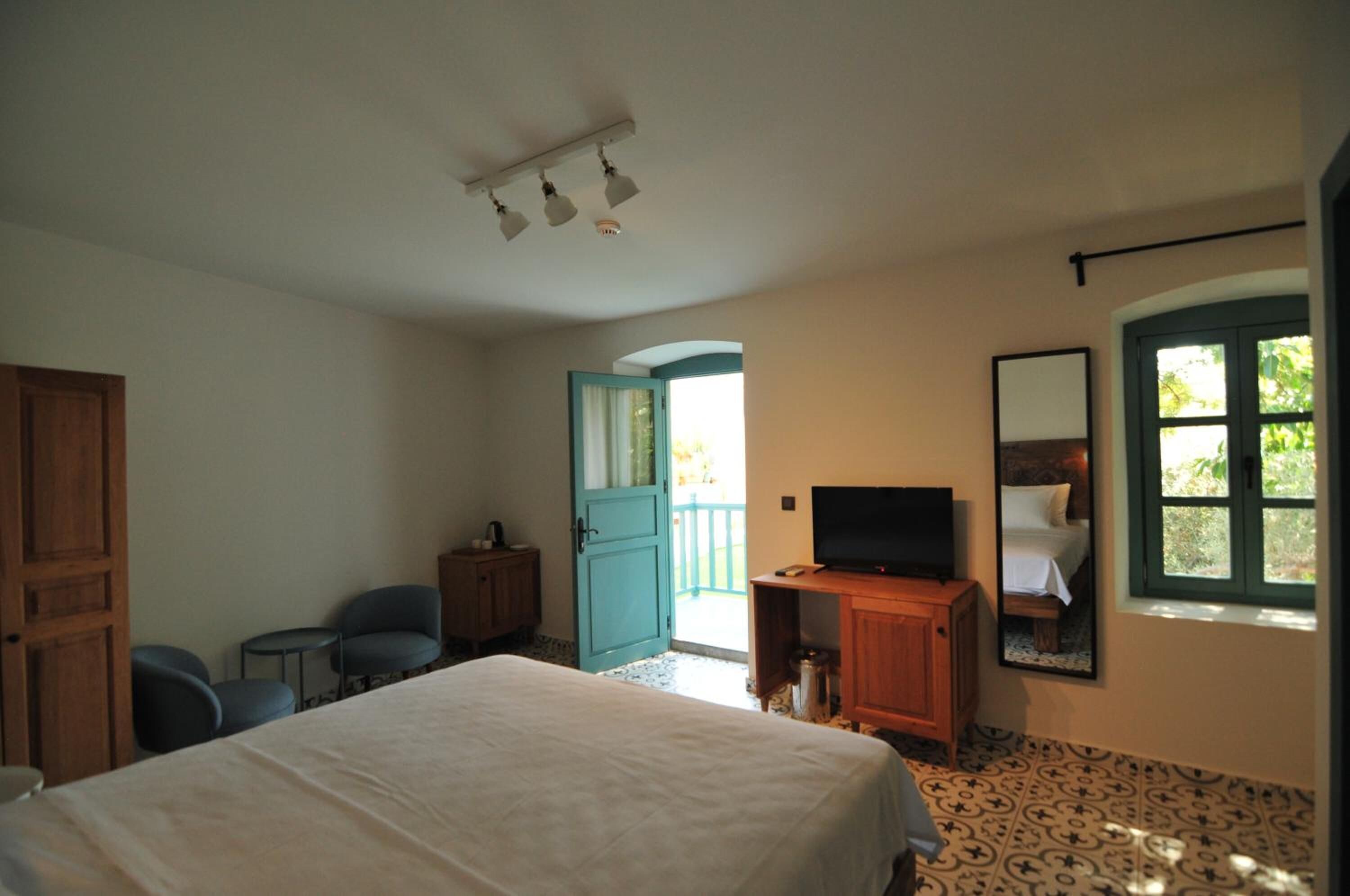 Un dormitorio con una cama, un televisor, un armario de madera, un escritorio, una silla, una puerta que da a un balcón y una ventana con vistas a la vegetación.