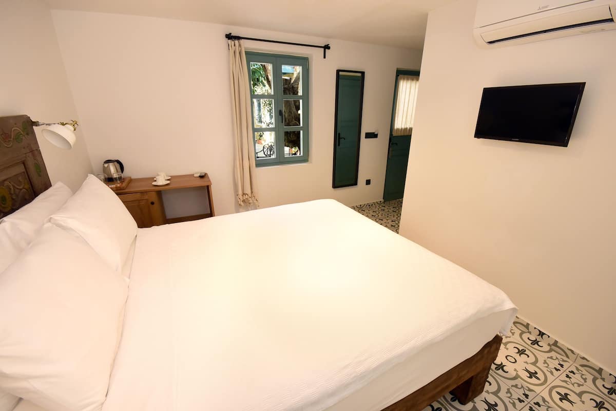 A neatly made bed with white linens, a bedside table with a cup and saucer, a window with curtains, a flat-screen TV mounted on the wall, and a patterned floor.