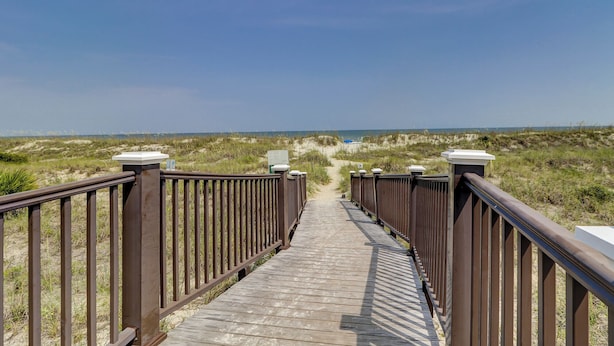 Ein hölzerner Steg führt zum Strand, umgeben von Dünen und blauem Himmel.