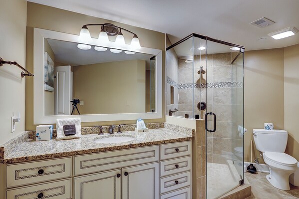 A bathroom with a large mirror, a vanity with a granite countertop, a glass-enclosed shower, and a toilet.