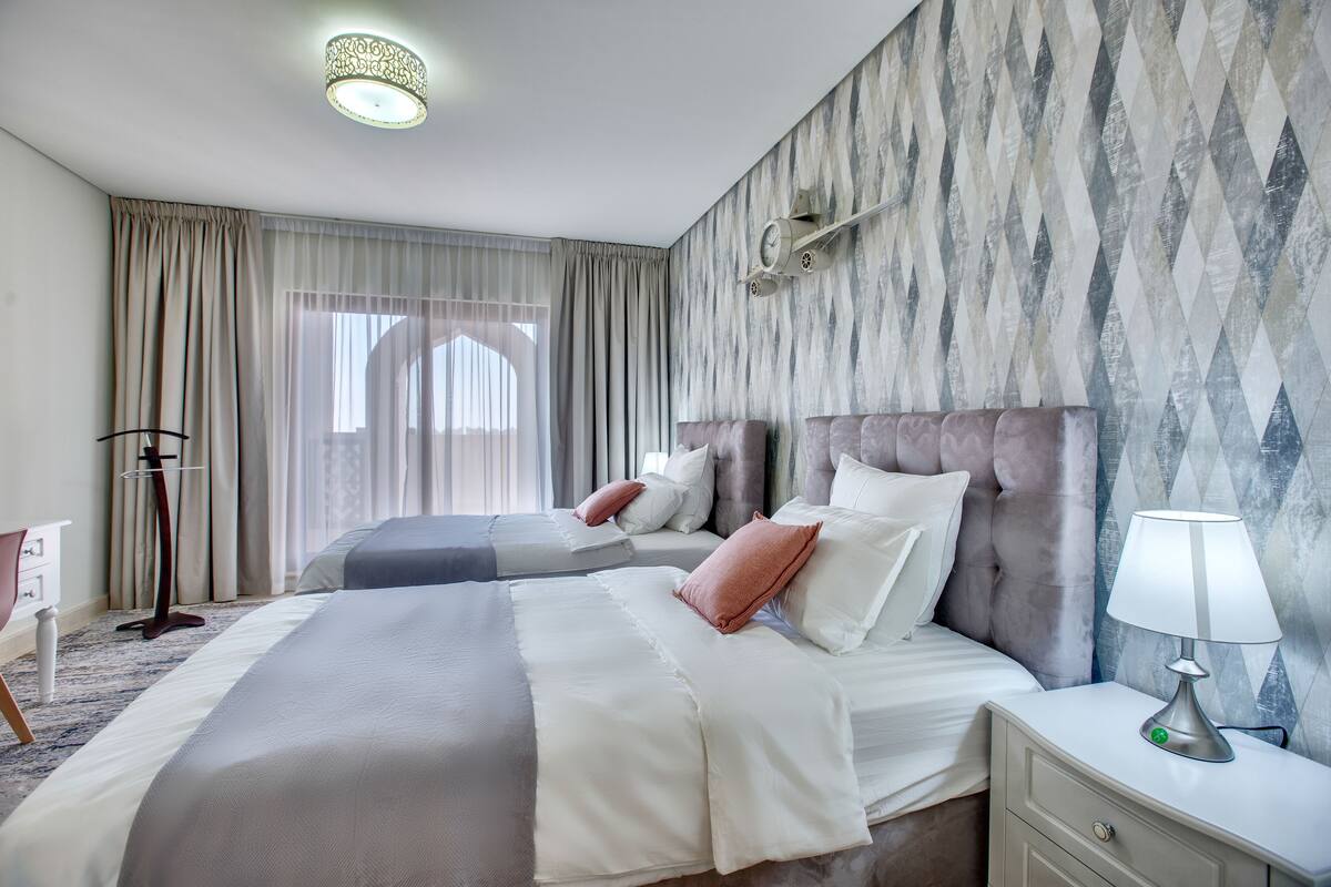 Une chambre d’hôtel avec deux lits, une table de chevet avec une lampe, un bureau et une grande fenêtre avec des rideaux.
