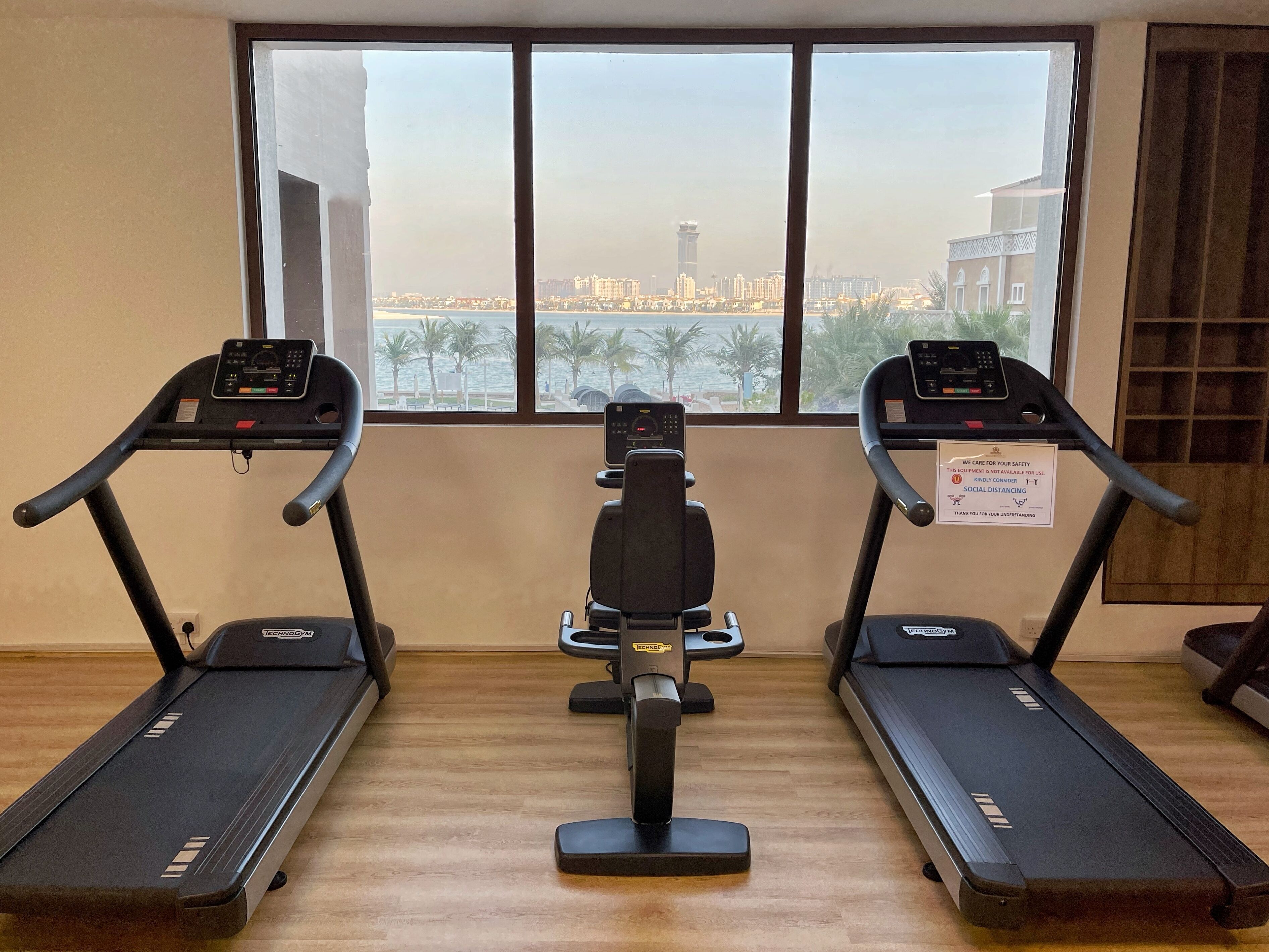 Academia com três esteiras, uma máquina elíptica central e uma grande janela com vista para a cidade.