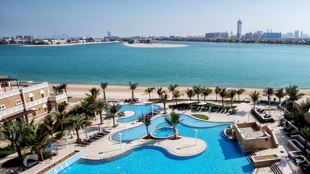 Una gran piscina de varios niveles con un diseño sinuoso, rodeada de palmeras y sillas de descanso, con una playa y el horizonte de la ciudad al fondo.