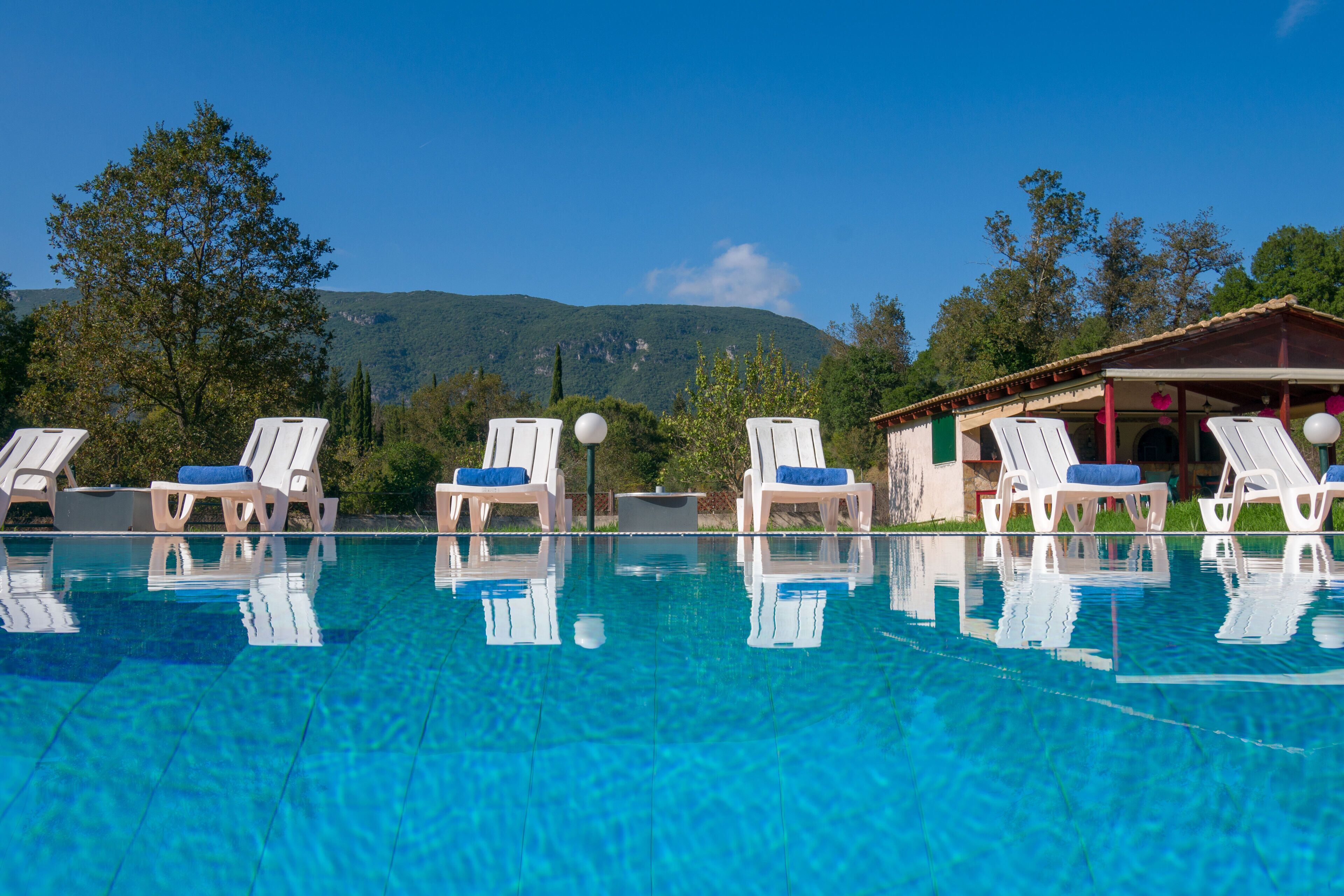 A clear swimming pool with white lounge chairs and blue cushions, surrounded by trees and a mountain in the background.