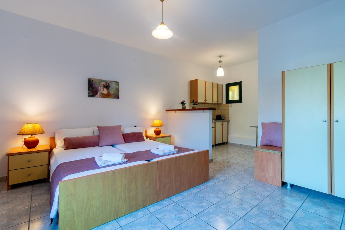 A modern bedroom with a large bed, bedside lamps, a painting on the wall, and a kitchenette area in the background.