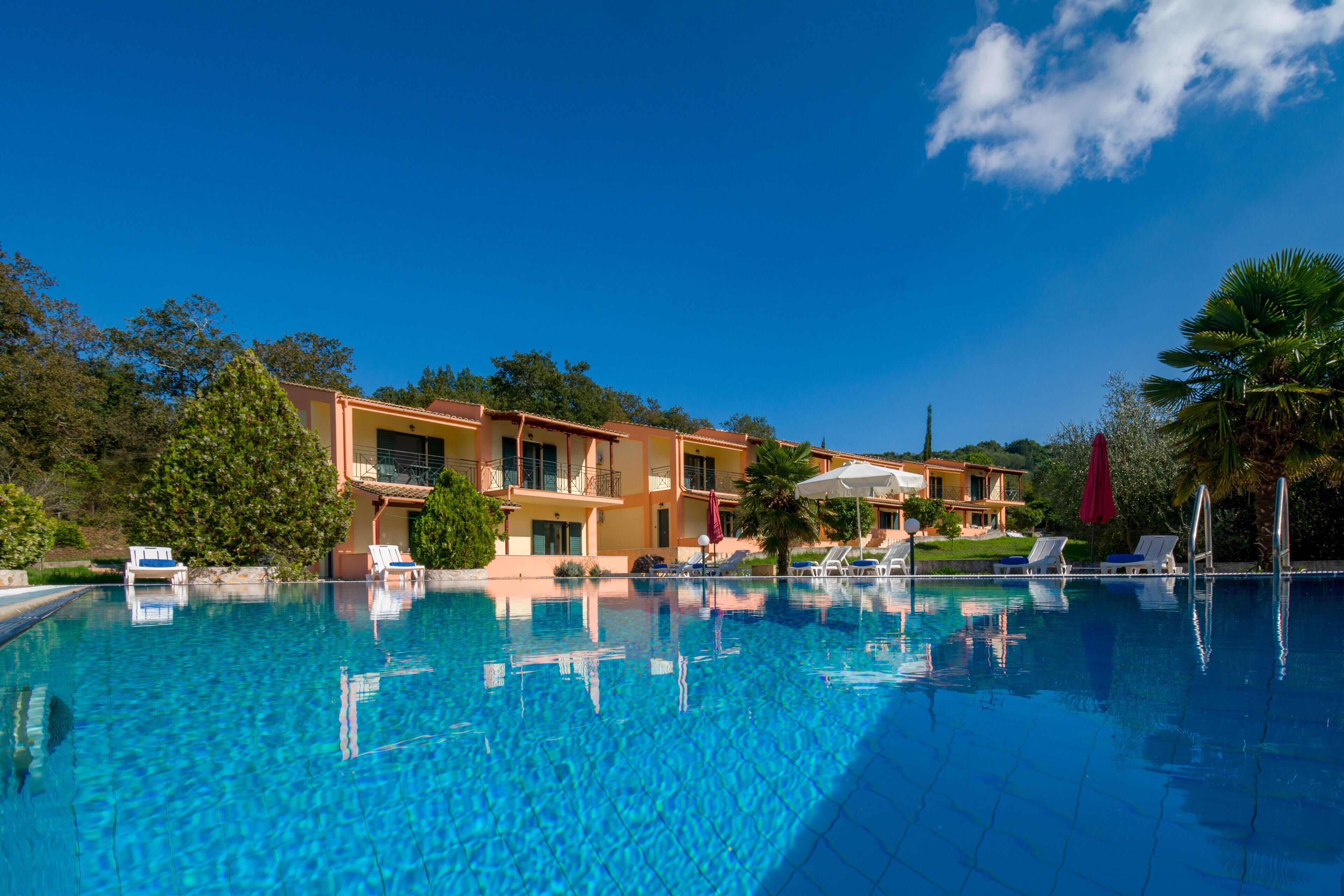 A swimming pool with lounge chairs and umbrellas, surrounded by a building with balconies and trees.