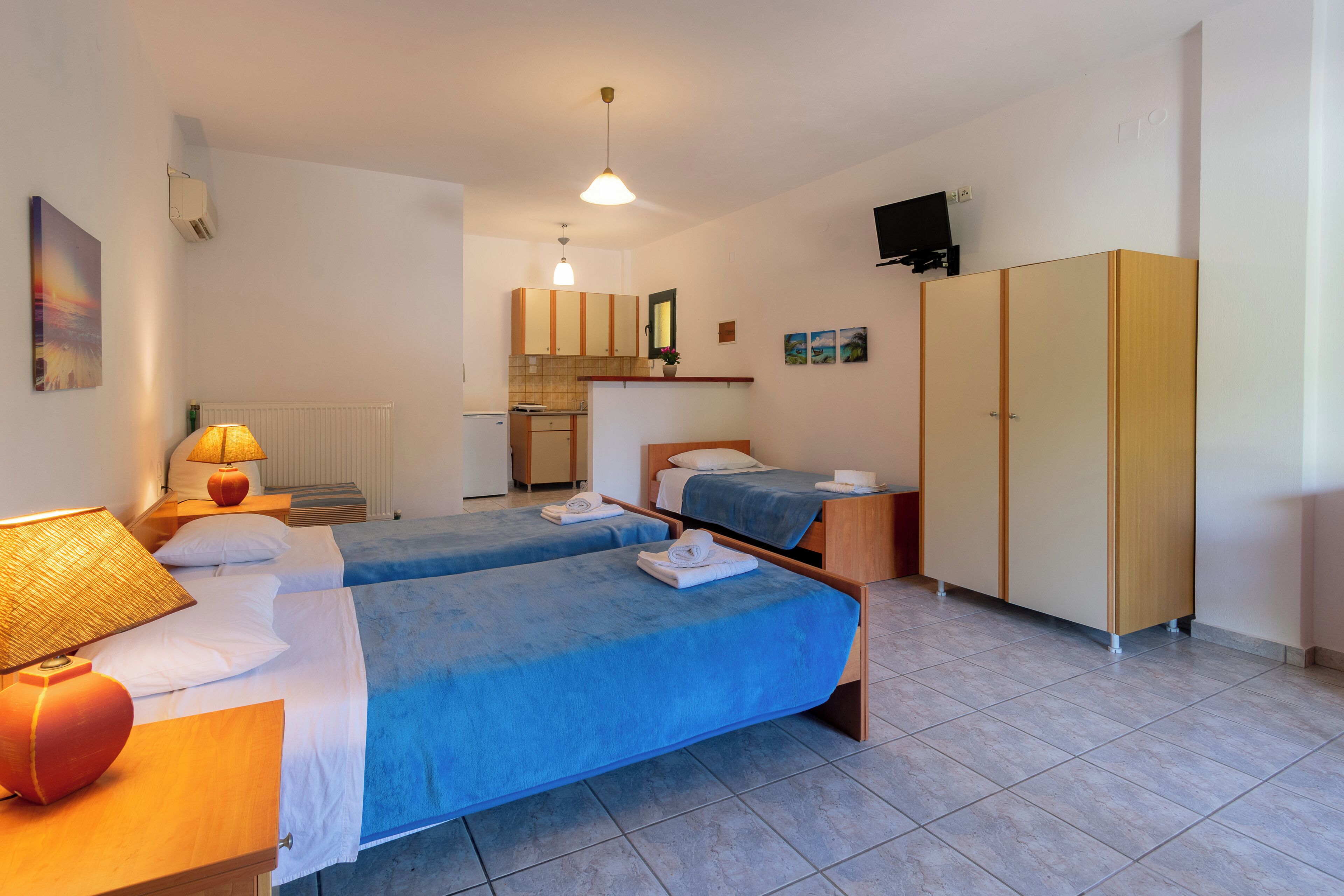 A hotel room with two beds, a kitchenette, a wardrobe, and a TV on the wall.
