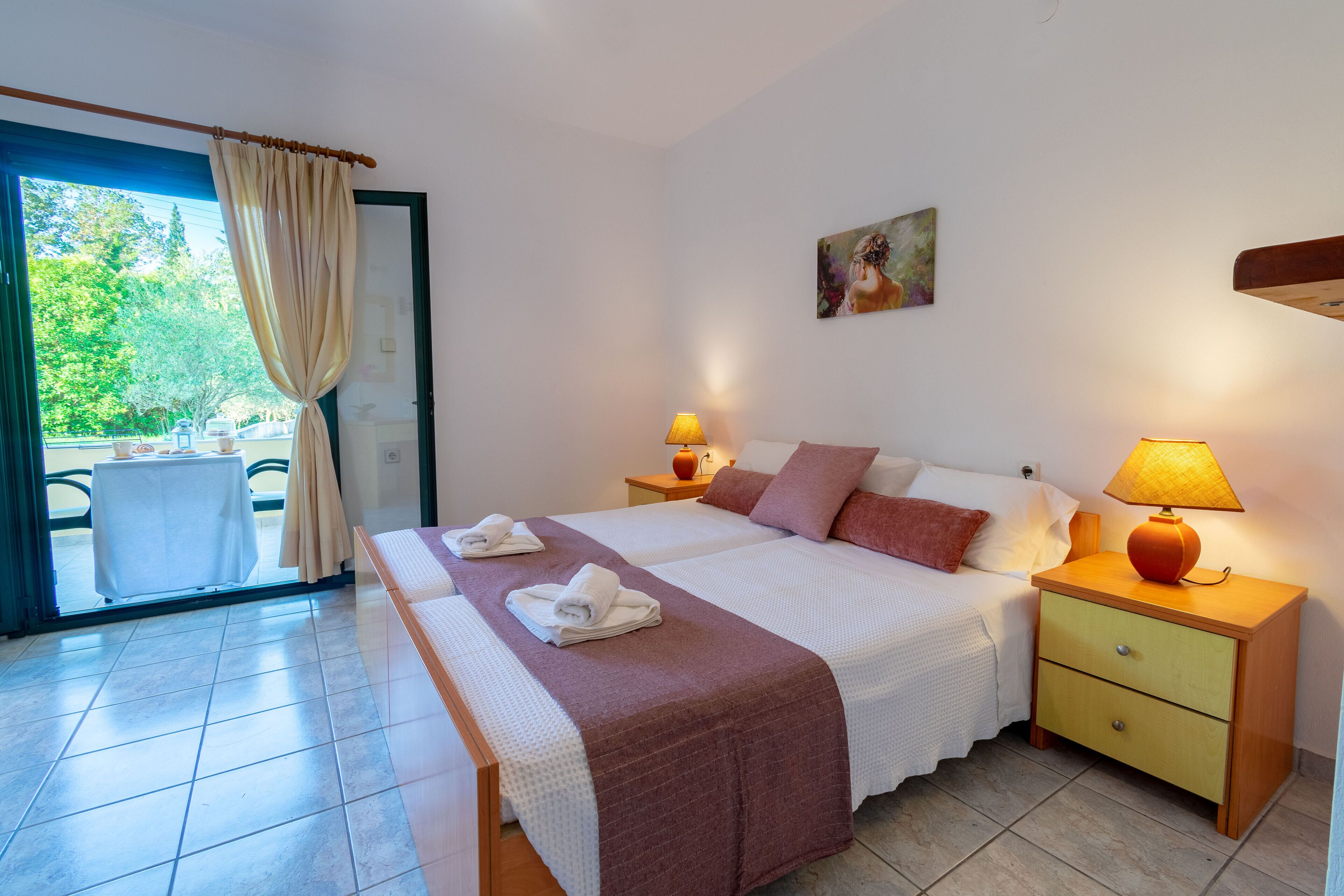 A hotel room with a bed, two bedside tables with lamps, a small table with a chair outside the door, and a painting on the wall.