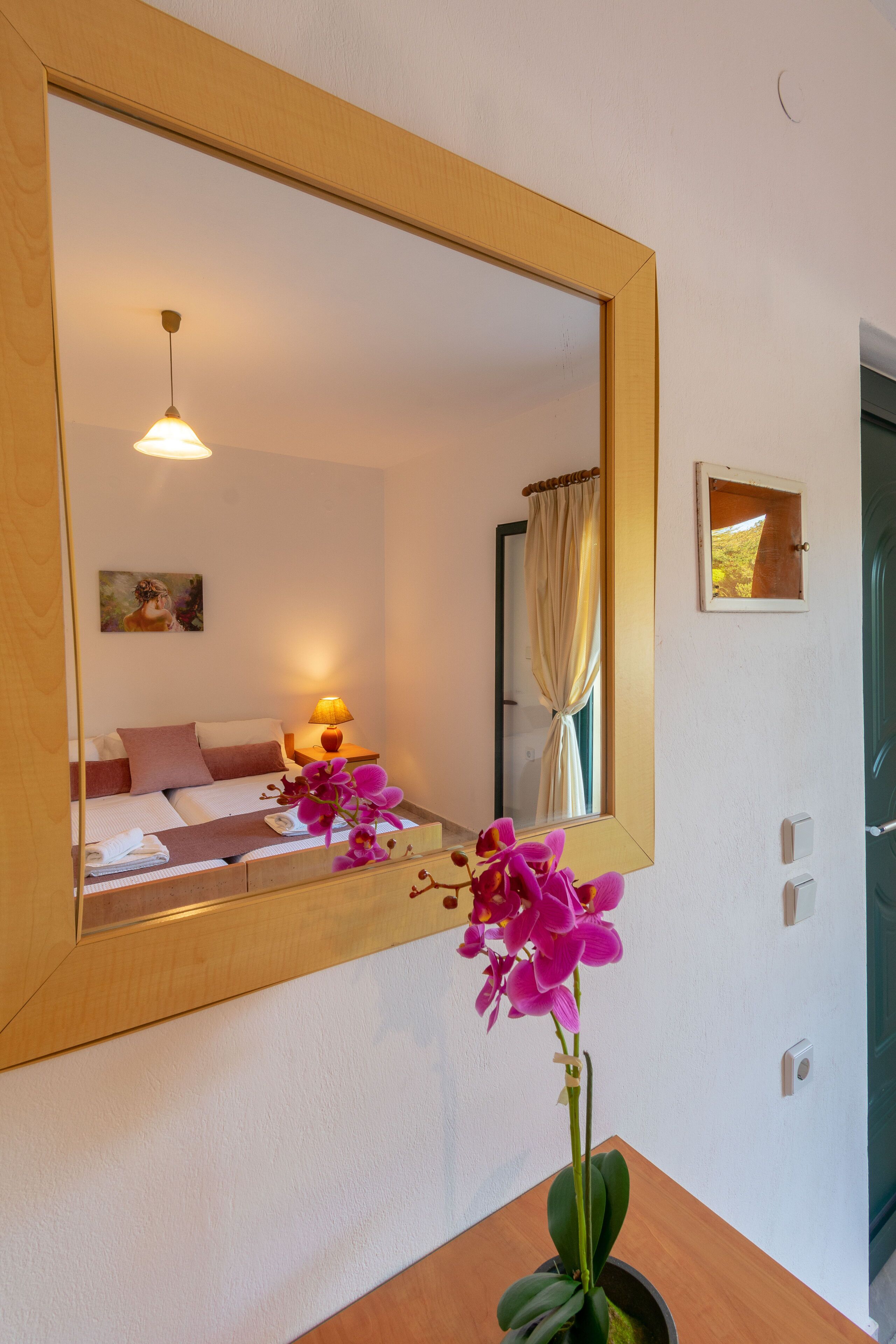 A bedroom with a large mirror, a bedside lamp, a bed with pillows, a small table with a vase of flowers, and a window with curtains.