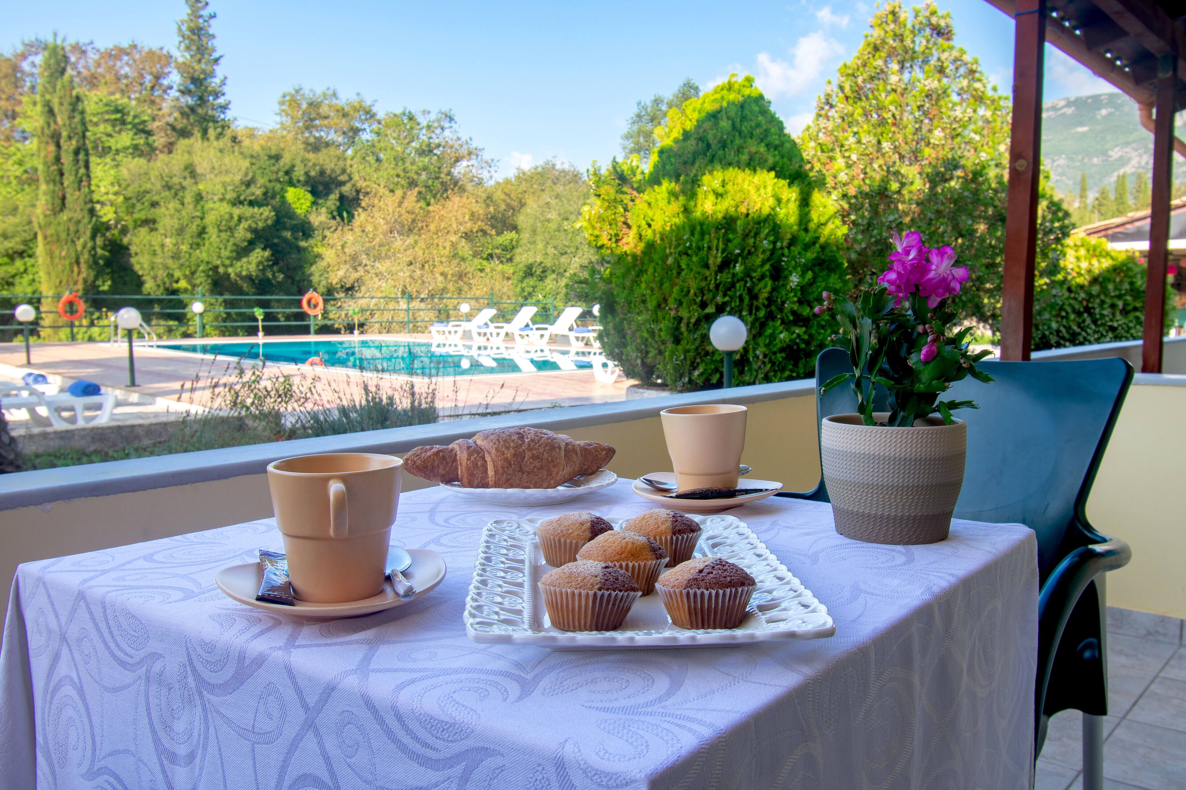 A table with a croissant, muffins, and a cup on a balcony overlooking a pool and greenery.