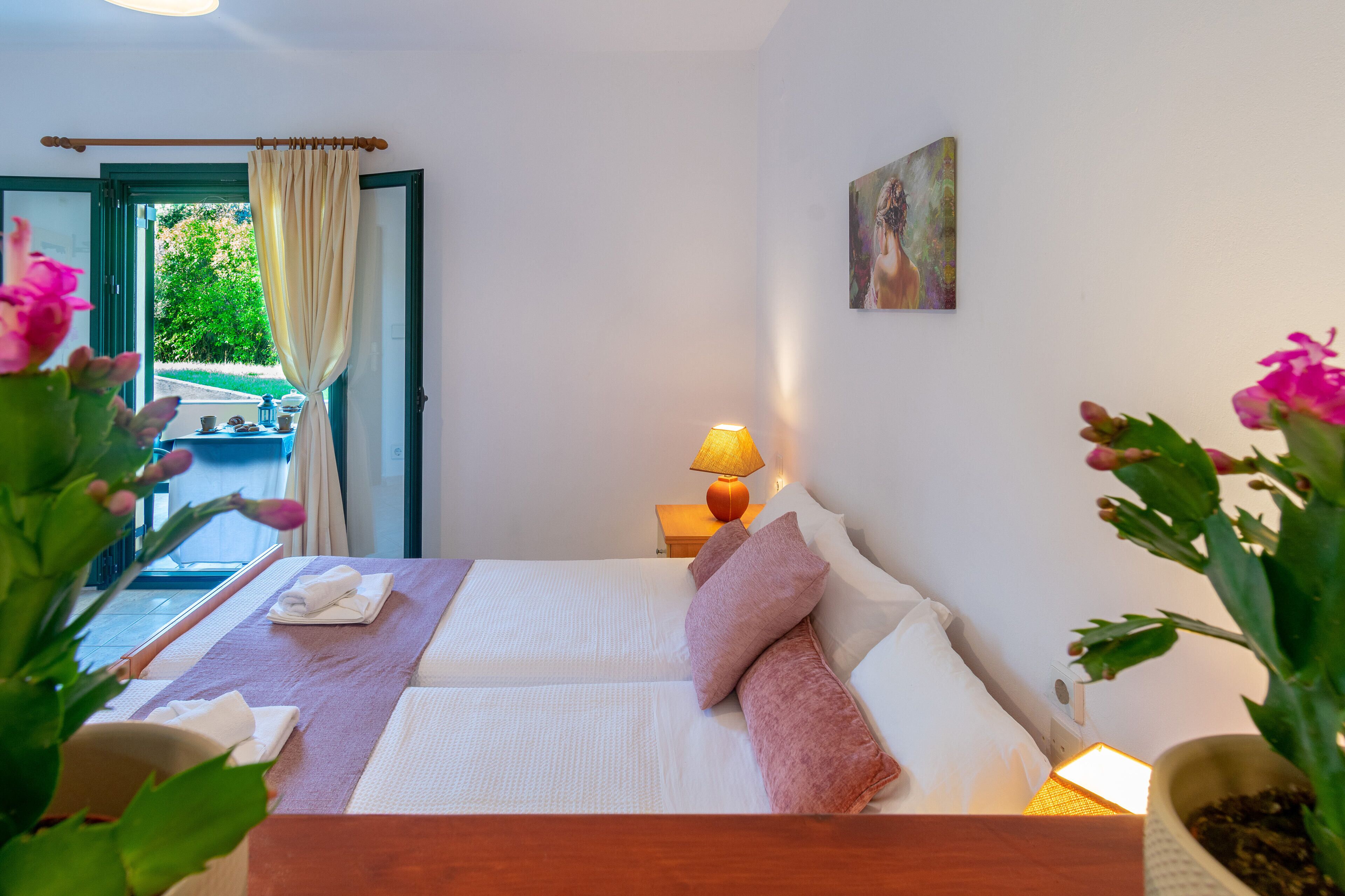 A hotel room with a bed, a bedside table with a lamp, a painting on the wall, and a view of the outdoors.