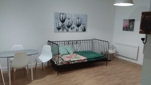Une petite pièce meublée, avec un canapé en métal noir, deux chaises blanches, une petite table ronde et un plaid vert.