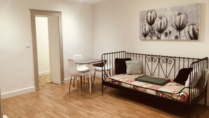 Une pièce avec un plancher en bois, un canapé en métal noir agrémenté de coussins à motifs floraux, une table blanche avec des chaises et un tableau accroché au mur.