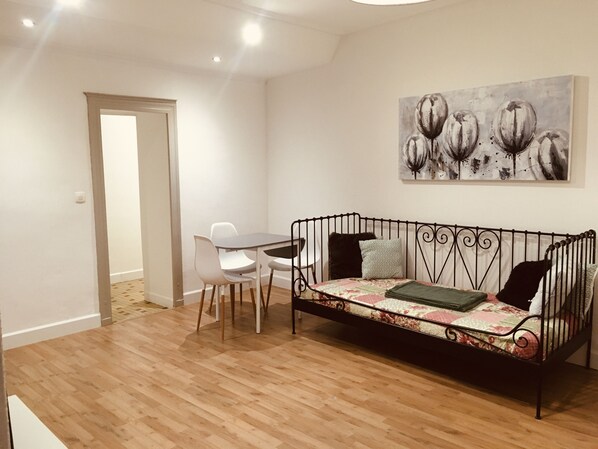 A room with a wooden floor, a black metal sofa with floral cushions, a white table with chairs, and a painting on the wall.