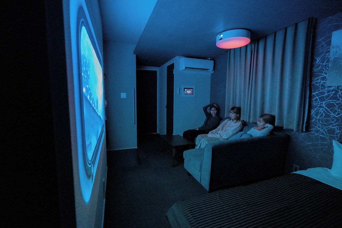 A dimly lit room with a large screen displaying a movie, a bed, and a wall-mounted air conditioner.