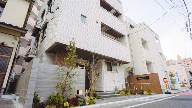 Un bâtiment moderne à la façade blanche, doté de nombreux balcons et d’un petit jardin agrémenté de plantes et d’un arbre.