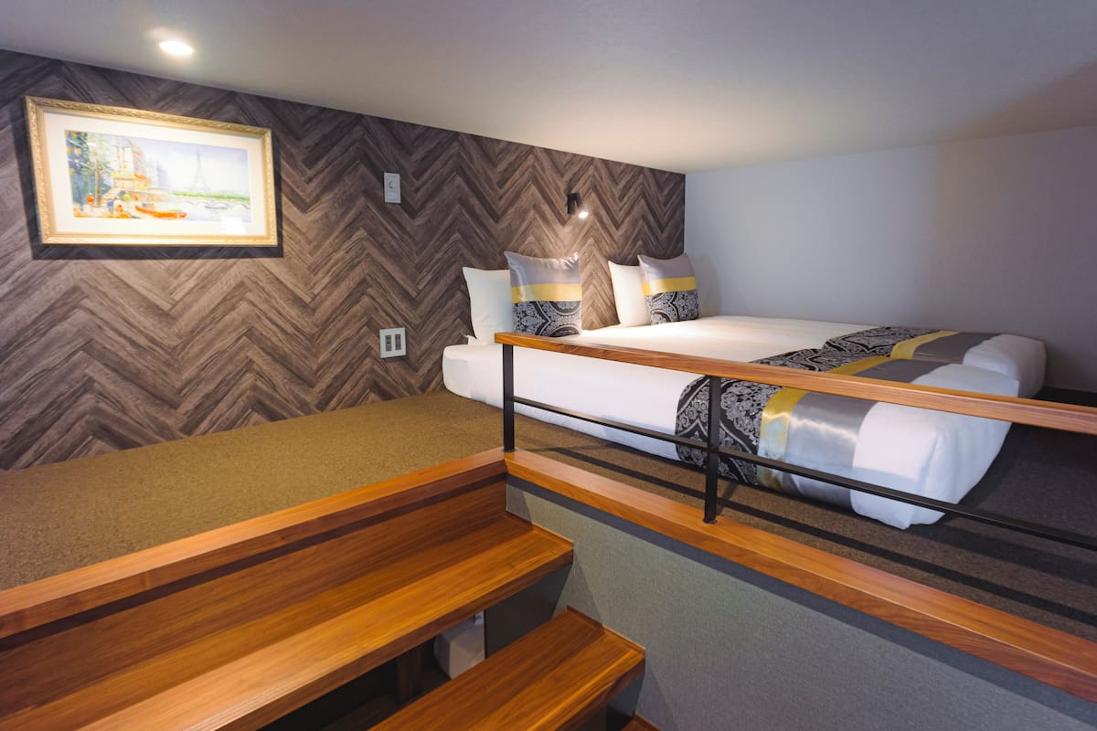 A bunk bed with a wooden frame and a lower bunk with a bench. There is a white bed with patterned pillows and a painting of the Eiffel Tower on the wall.