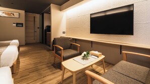 Ein modernes Hotelzimmer mit Holzboden, einem Flachbildfernseher an der Wand, einer Couch, einem kleinen Tisch mit einem dekorativen Gegenstand und einem Sessel.