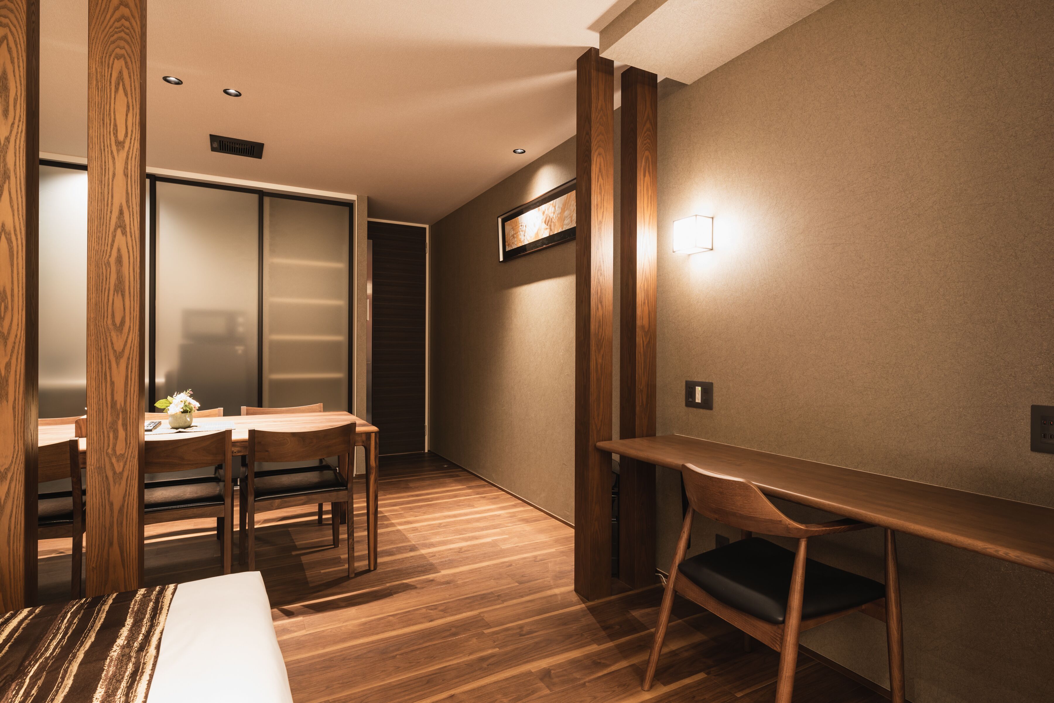 A hotel room with a wooden floor, a dining table with chairs, a bed, a sliding door, and a wall-mounted light fixture.