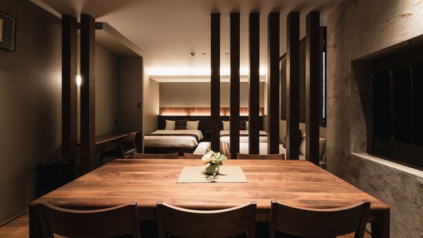Ein geräumiger Essbereich mit Holztisch und -stühlen, einer kleinen Vase mit Blumen und Blick auf ein Schlafzimmer mit Bett und Nachttisch.