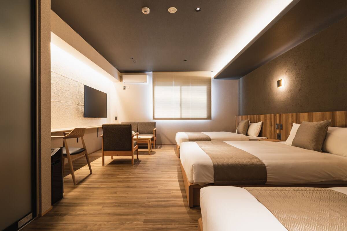 Quarto de hotel moderno com chão de madeira, duas camas, uma escrivaninha com cadeira, televisão fixada na parede e janela com persianas.