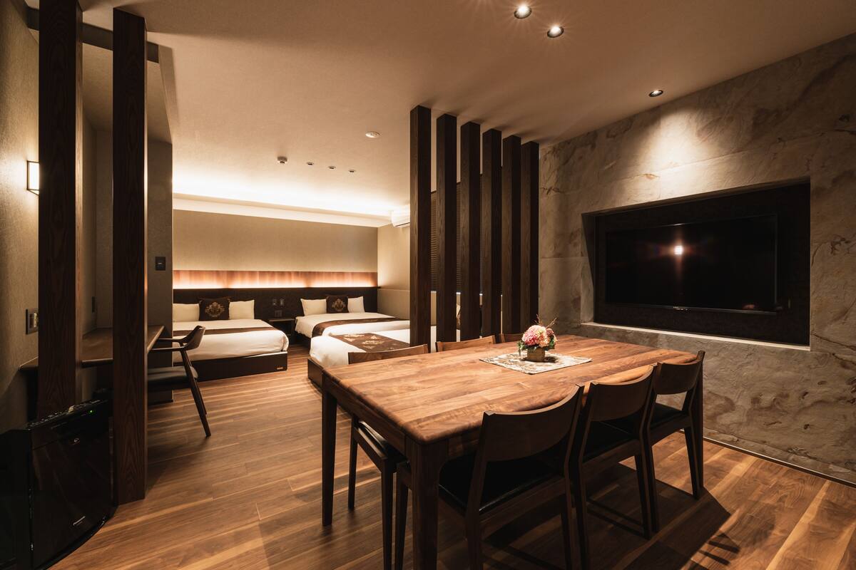 Quarto de hotel moderno com uma mesa de jantar de madeira, duas camas, uma televisão e uma janela grande.