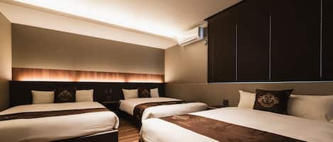 A hotel room with three beds, a wooden floor, a large window with blinds, and a wall-mounted air conditioning unit.