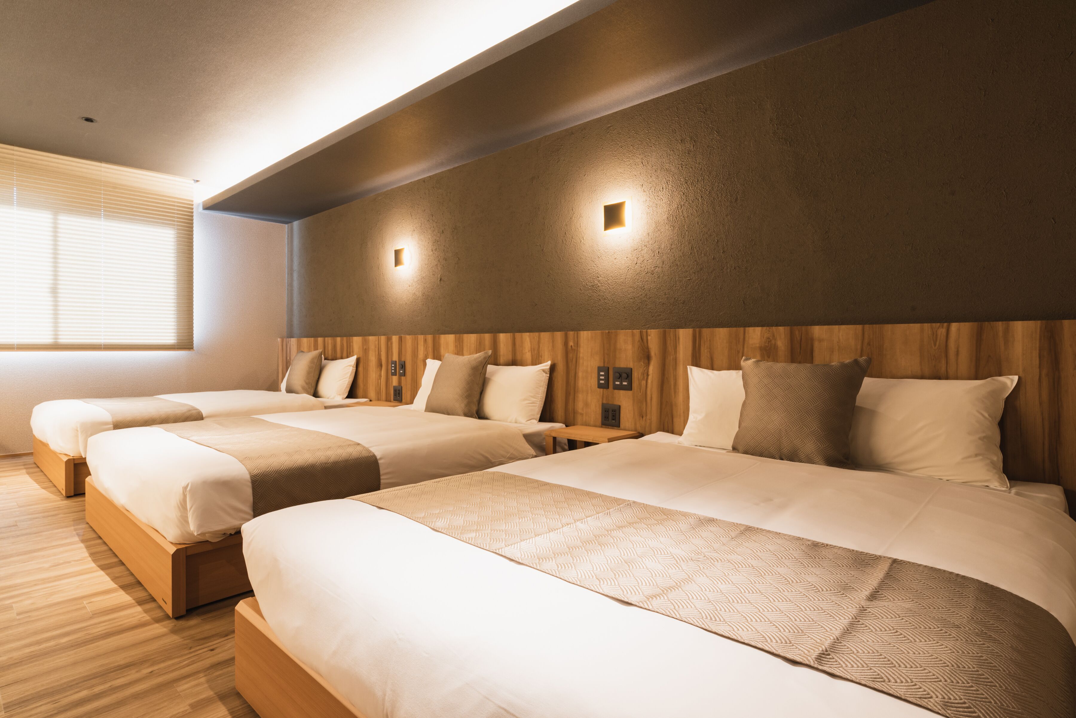 A hotel room with three beds arranged in a row, wooden headboard, and a large window with blinds.