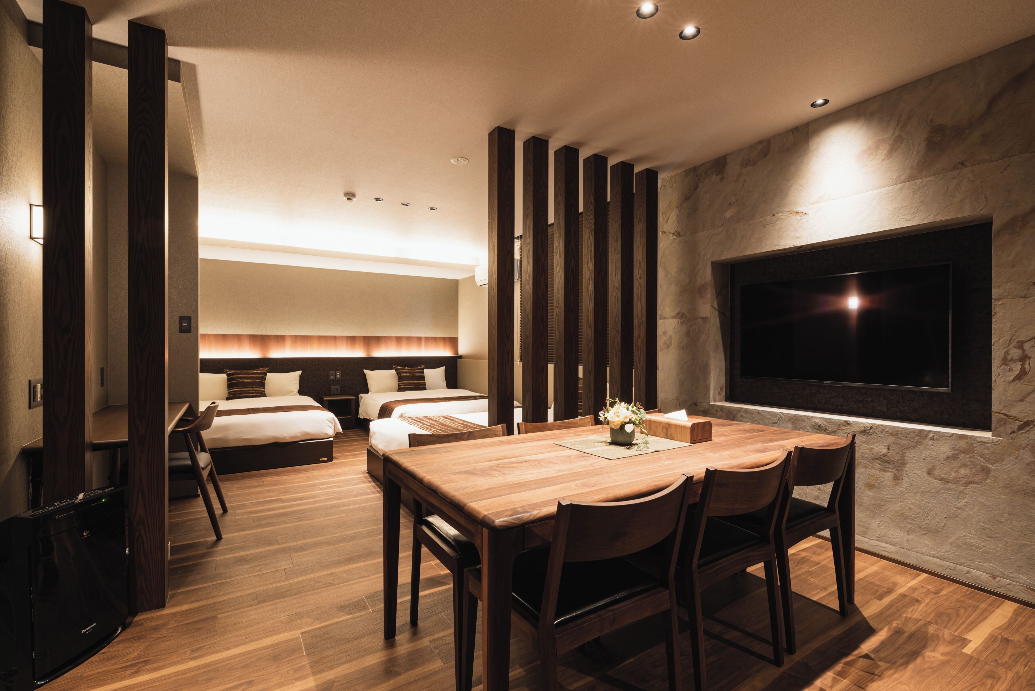 A modern hotel room with a wooden dining table, two beds, a TV, and a stone wall.