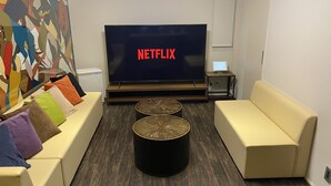A modern living room with a large flat-screen TV displaying 'NETFLIX', a sofa, a coffee table, and a small side table with a laptop. - Bond Base (Fukuoka)
