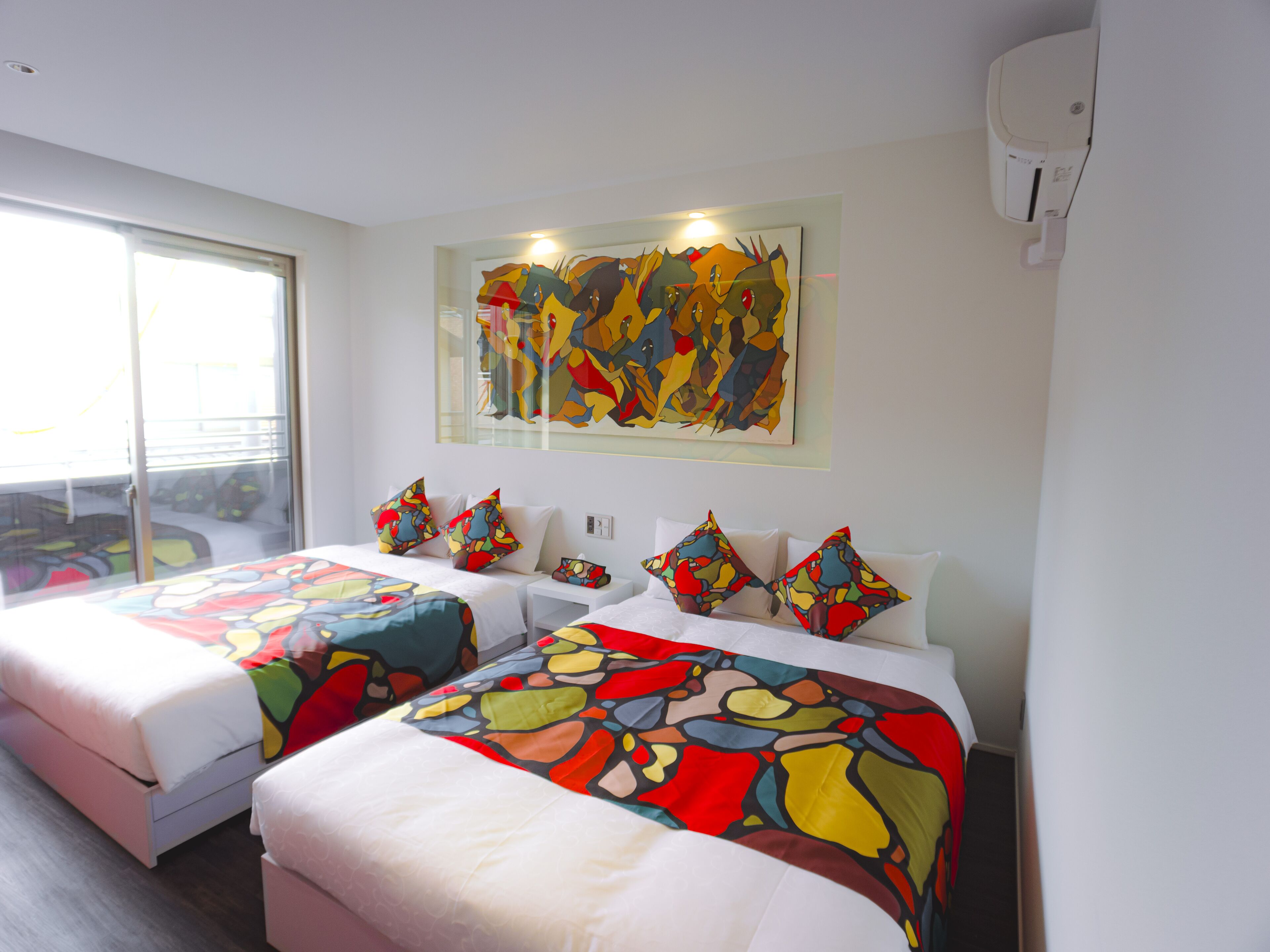 Ein Hotelzimmer mit zwei Betten, farbenfroher Wandkunst und einem großen Fenster.