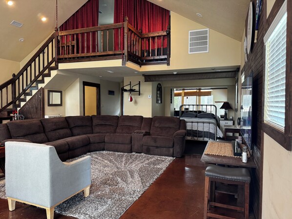 A spacious living area with a sectional sofa, a small armchair, and a wooden coffee table. A staircase leads to an upper level with red curtains.